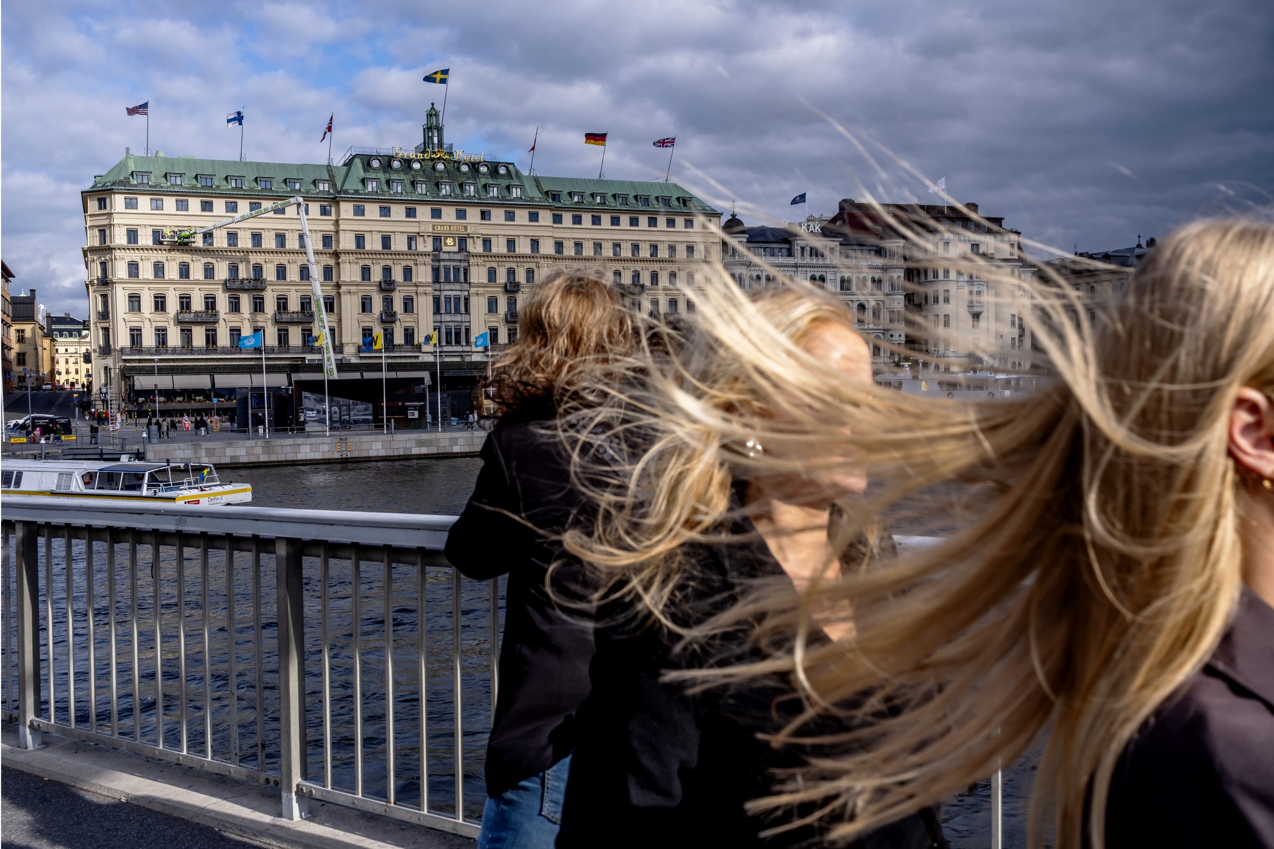Vy över Grand Hôtel i centrala Stockholm med svenska och internationella flaggor på taket. I förgrunden syns människor med vinden i håret, vilket skapar rörelse i bilden. Himlen är mulen, men byggnaden är belyst av dagsljus.