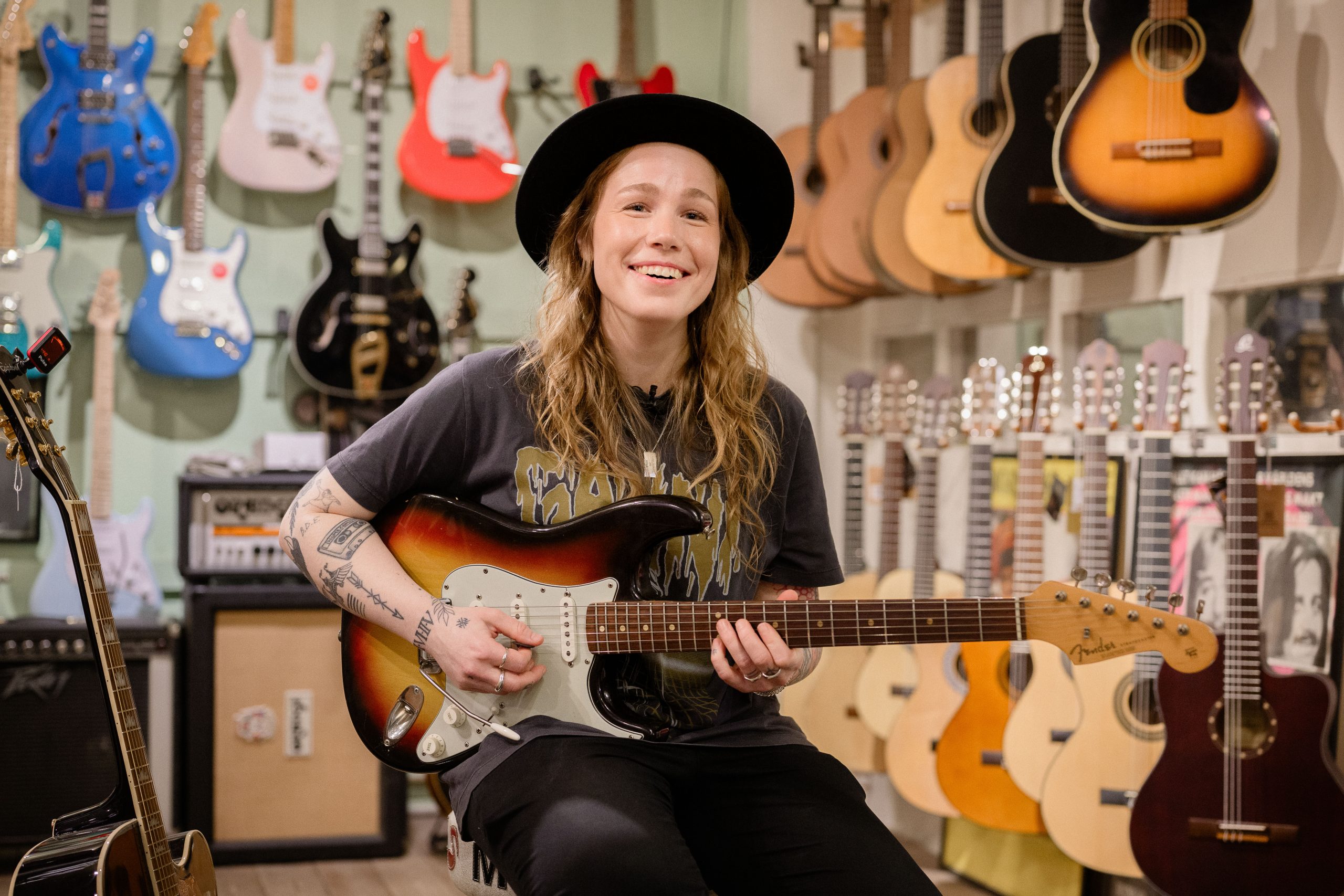 Rebecca Mardal, kompositör och gitarrist, sitter i en gitarrbutik och spelar elgitarr. Hon har t-shirt med tryck och hatt och ler.