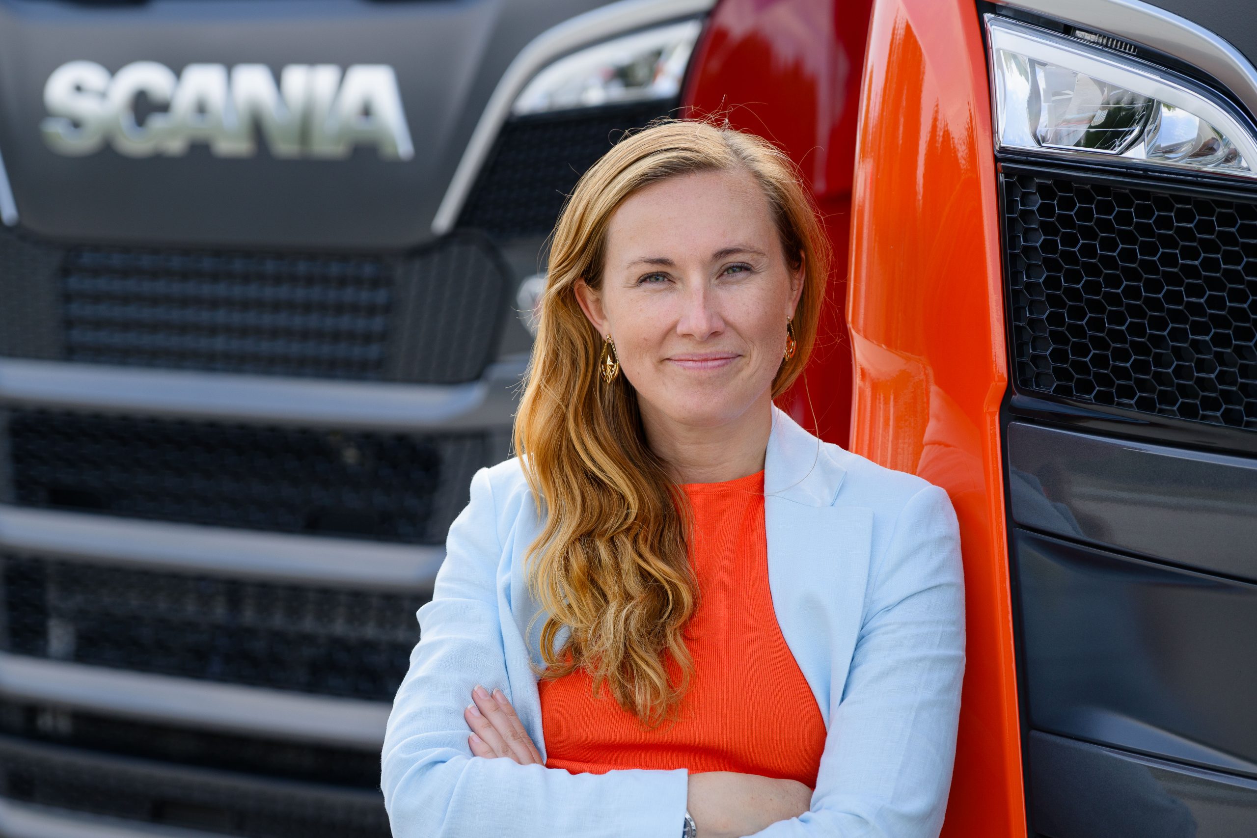 Sara Hermansson, Head of Power Solutions, Scania Group, står vid en orange lastbil. Hon har rött långt hår, orange topp och en ljusblå kavaj och ler.