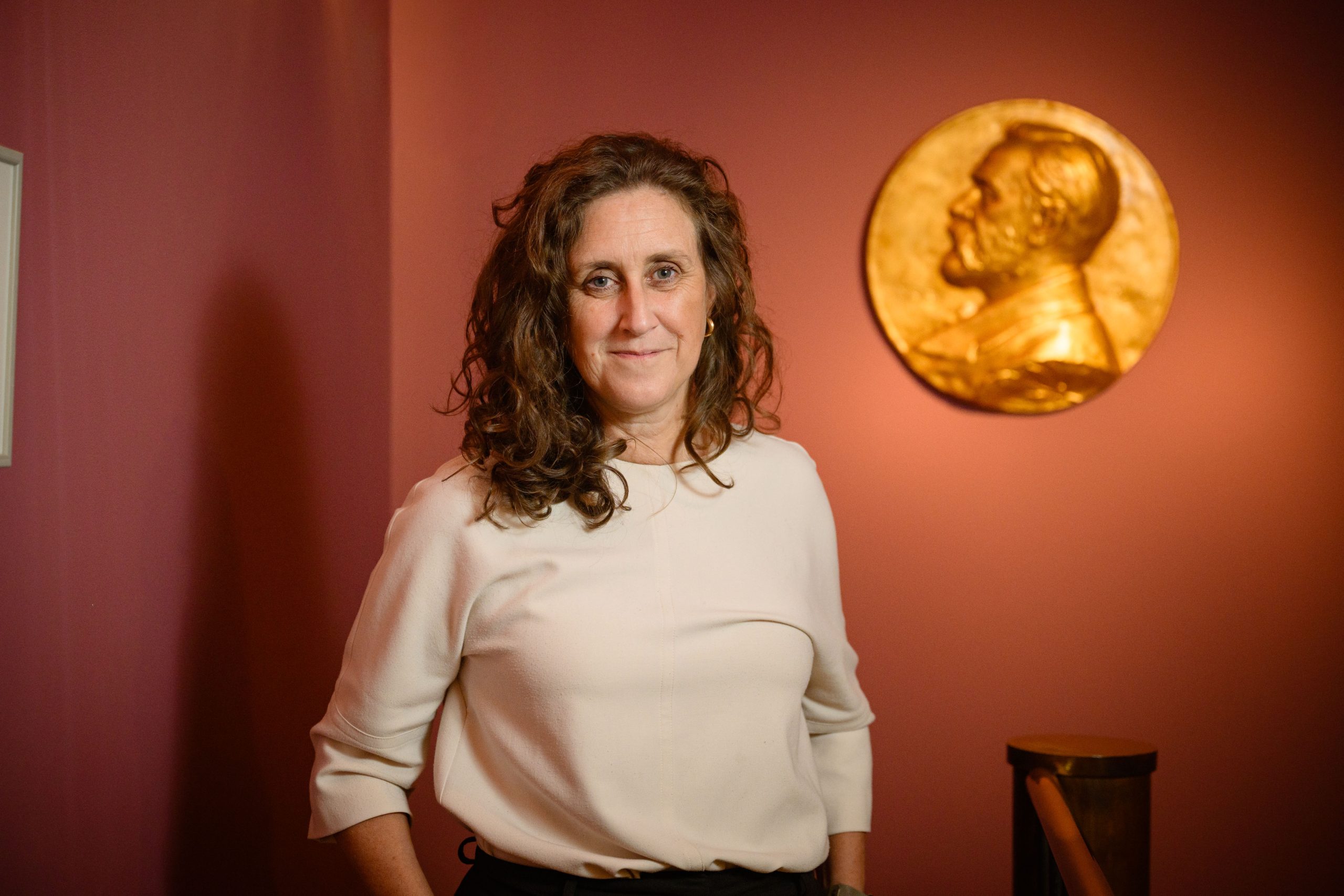 Hanna Stjärne, vd för Nobelstiftelsen, står framför en vägg i rosa nyans med en stor Nobelmedalj i guld upplyst i bakgrunden. Hon bär en ljus tröja och ler in i kameran.