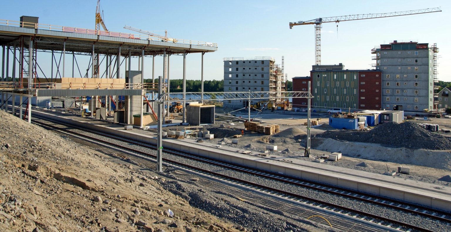 Byggarbetsplats för järnväg med spår, perrong och bostadshus under uppförande i stadsmiljö