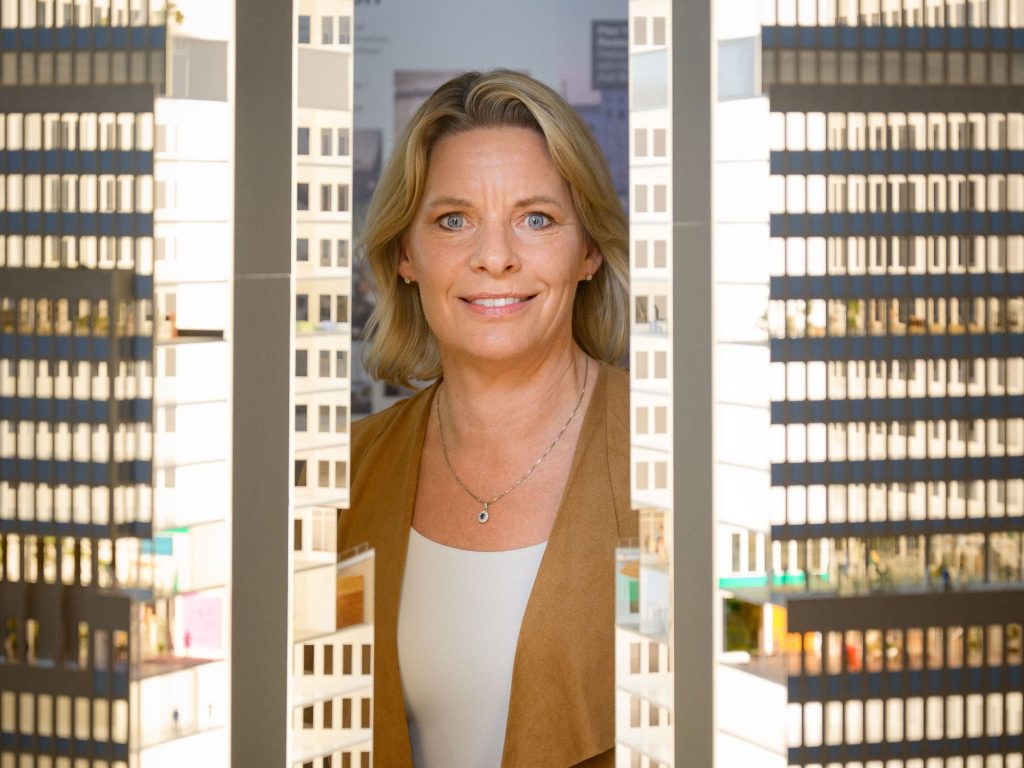 Annika Ånäs, vd för Atrium Ljungberg, står leende mellan två höga arkitekturmodeller av kontorshus. Bilden är tagen inomhus i ett upplyst rum med fokus på framtida stadsutveckling.