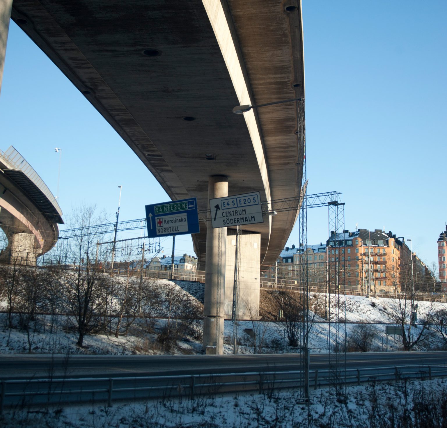 Motorväg och järnväg som möts under broar i stadsmiljö med vägskyltar mot E4 och centrala Stockholm
