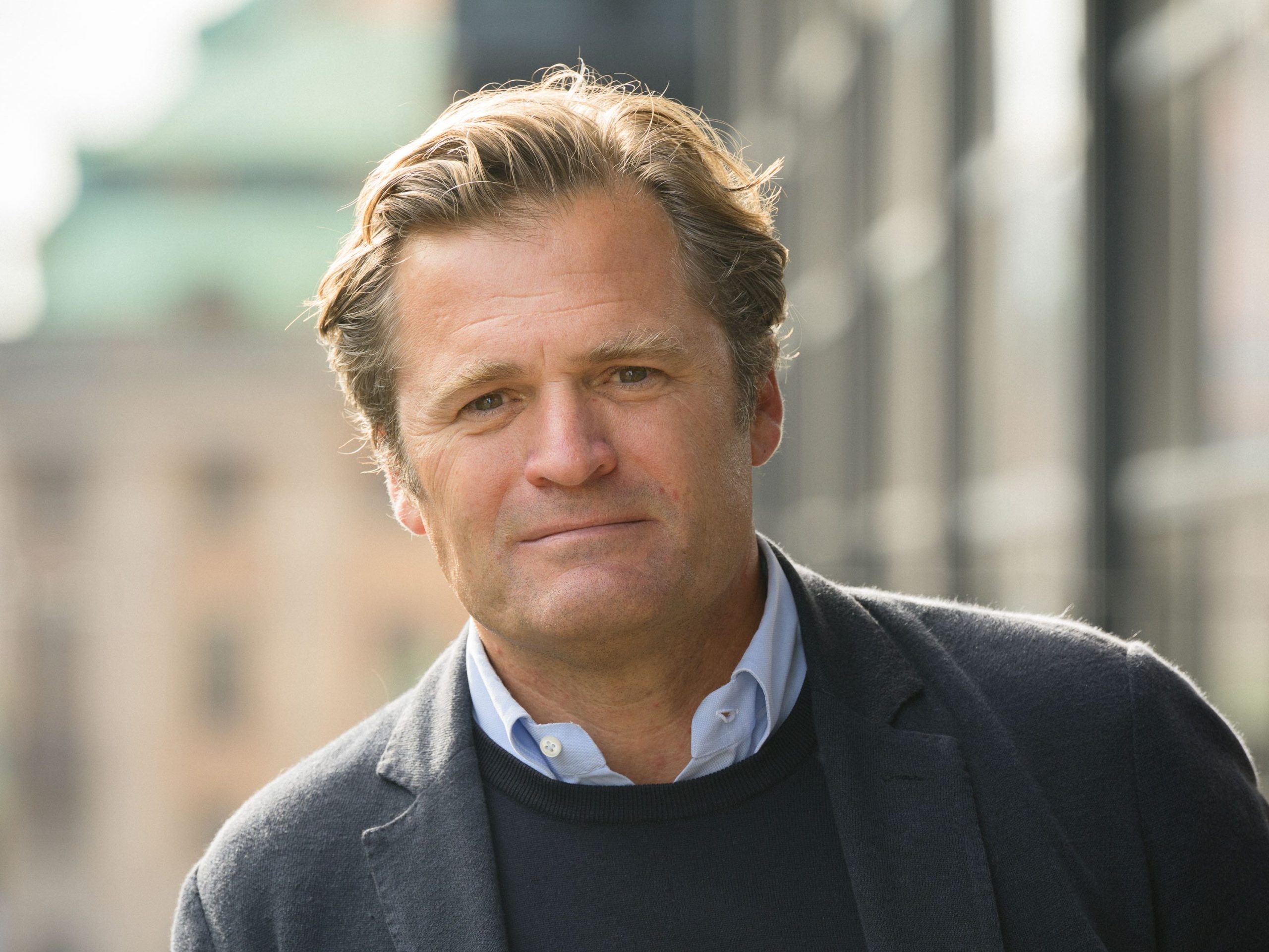 Adam Kostyal, vd på Nasdaq, står utomhus lutad mot ett räcke med oskarpa byggnader i bakgrunden. Han är klädd i mörk kavaj och tittar in i kameran med ett allvarligt uttryck.